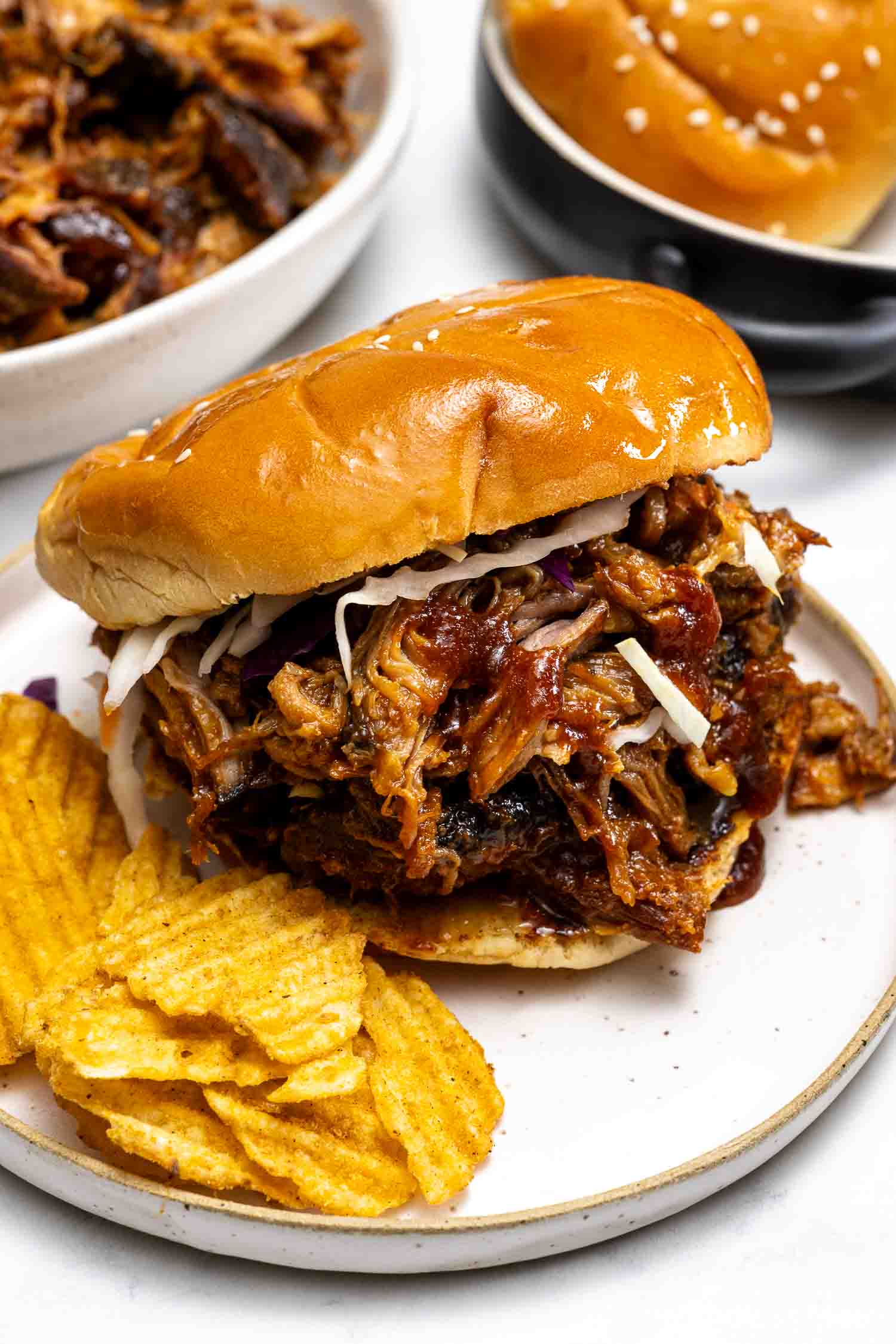 Hot Honey Pulled Pork sandwich with barbecue sauce and coleslaw on a bun, served with ridged potato chips on a white plate.