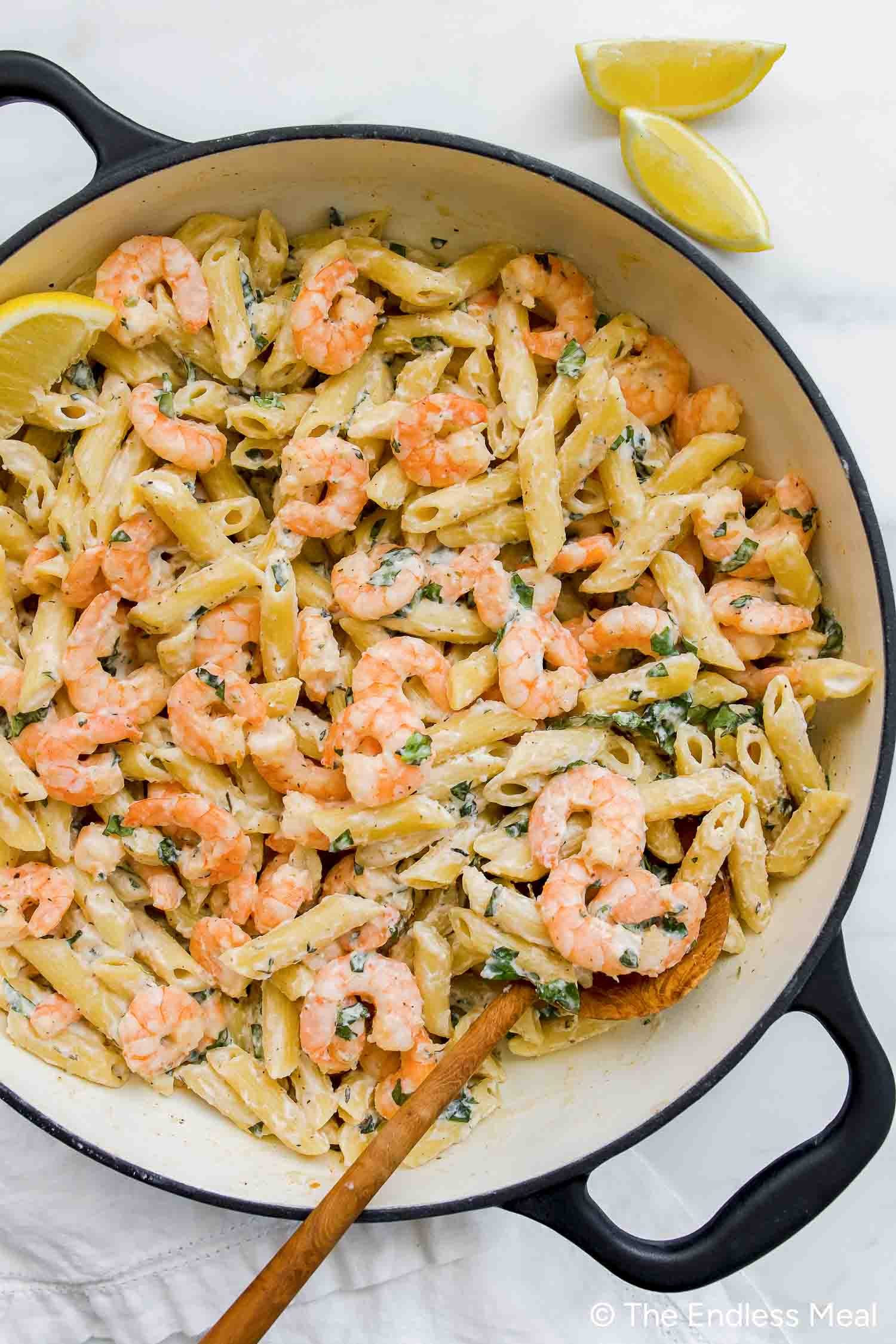 Cooked penne pasta and shrimp tossed in a lemony ricotta pasta sauce, garnished with herbs and lemon wedges in a large pan, with a wooden spoon resting inside.
