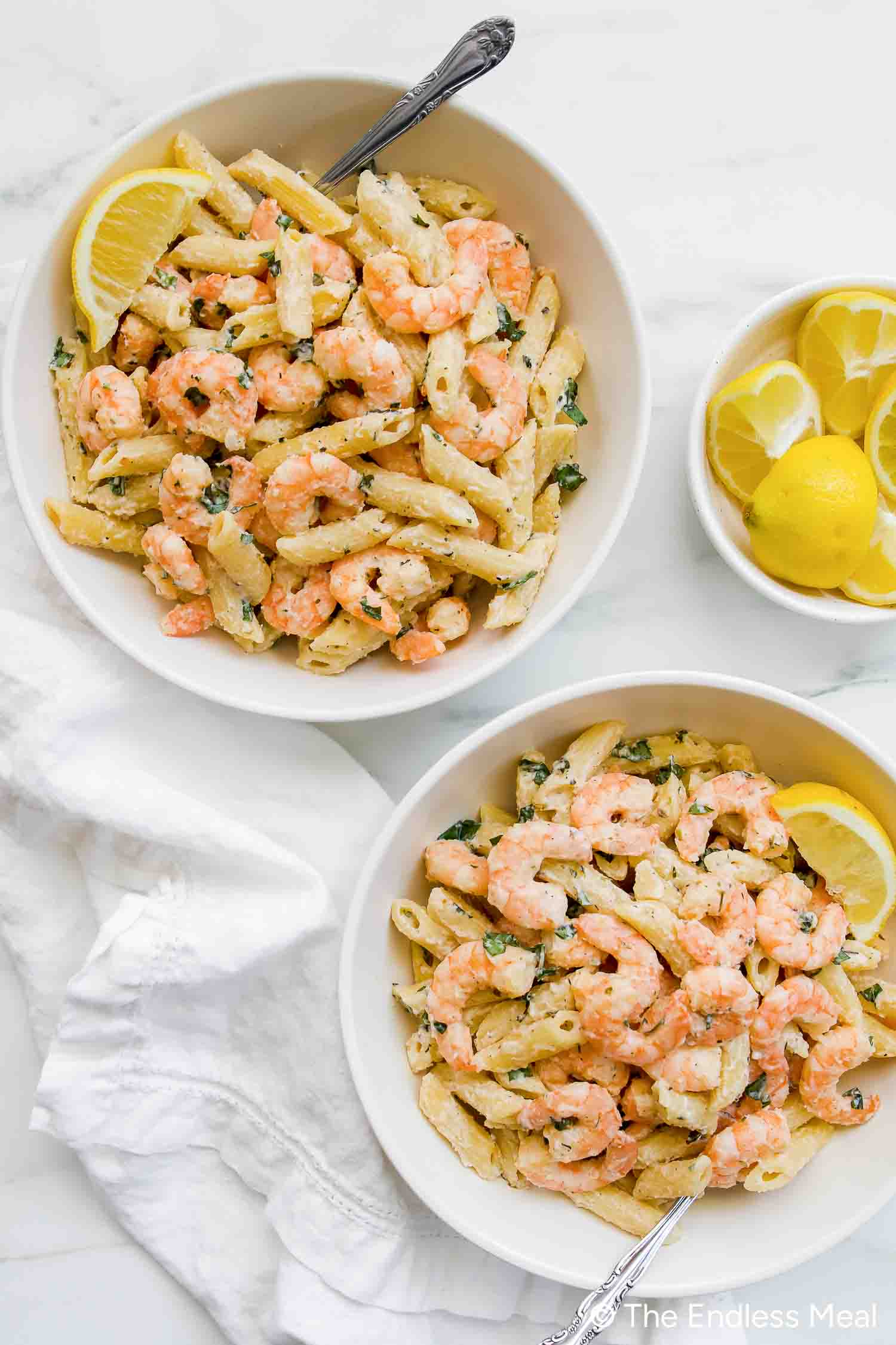 Two bowls of penne pasta with shrimp in a creamy, lemony ricotta pasta sauce, garnished with lemon wedges, sit beside a small bowl of sliced lemons on a white surface.