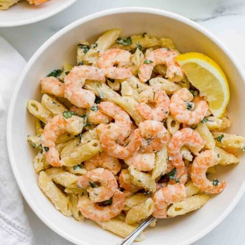 A bowl of shrimp pasta mixed with penne and herbs, topped with a lemon wedge and a fork resting inside, features a light lemony pasta sauce for a fresh, zesty finish.