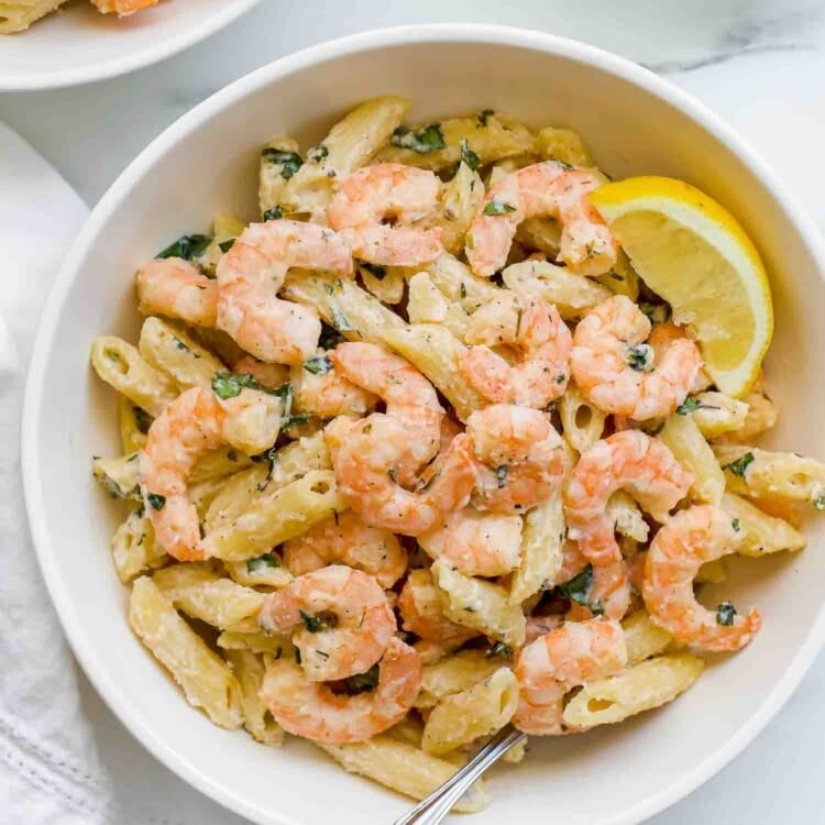 A bowl of shrimp pasta mixed with penne and herbs, topped with a lemon wedge and a fork resting inside, features a light lemony pasta sauce for a fresh, zesty finish.