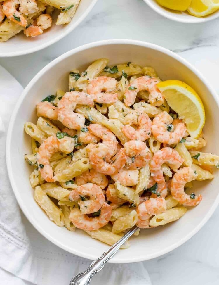 A bowl of shrimp pasta mixed with penne and herbs, topped with a lemon wedge and a fork resting inside, features a light lemony pasta sauce for a fresh, zesty finish.
