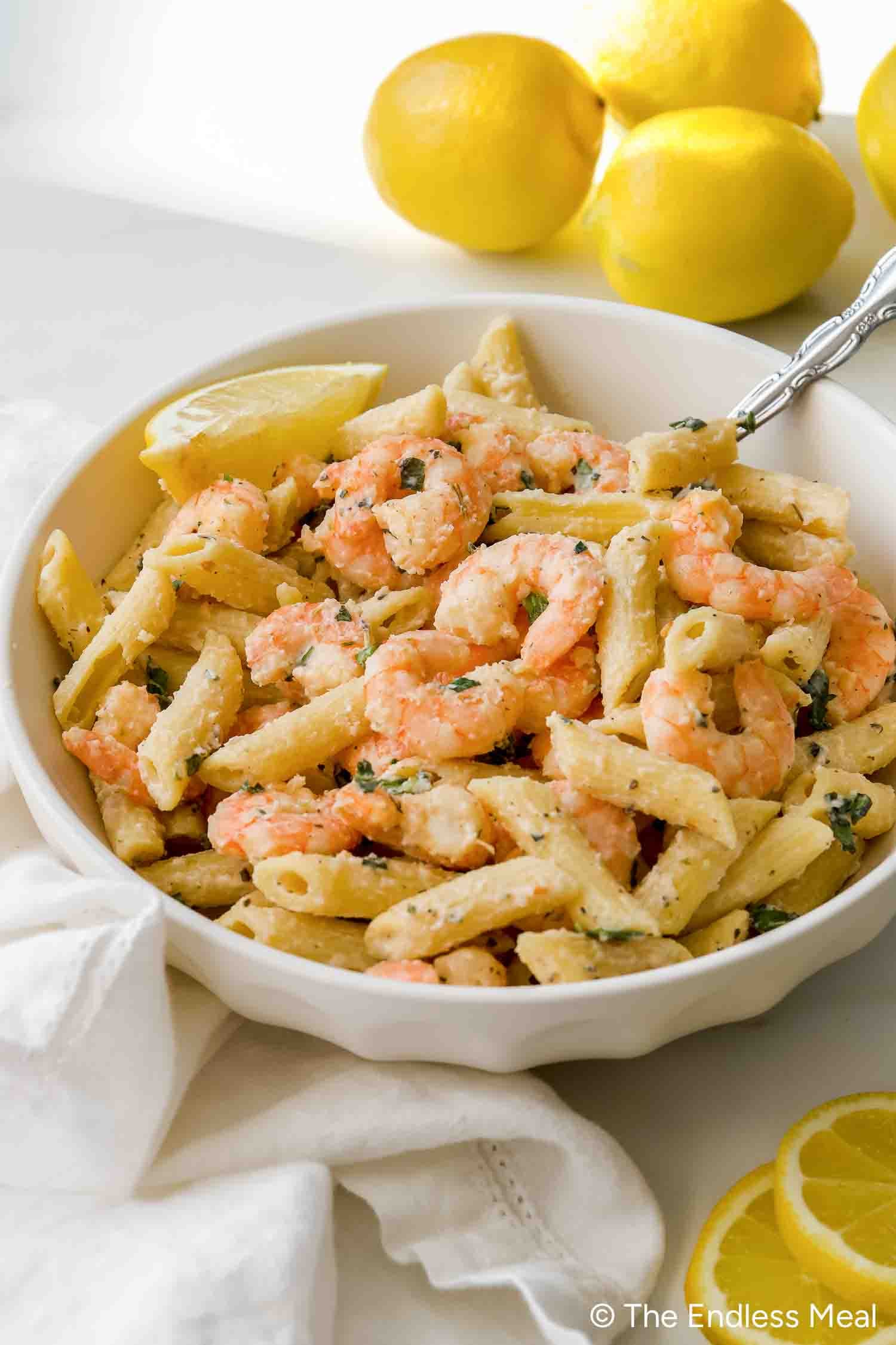 A bowl of penne pasta with shrimp in a creamy, lemony ricotta pasta sauce, garnished with herbs and a lemon wedge, with whole lemons in the background.
