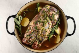 A seasoned Braised Leg of Lamb sits in a Dutch oven with fresh herbs, onions, and broth, ready to be cooked.
