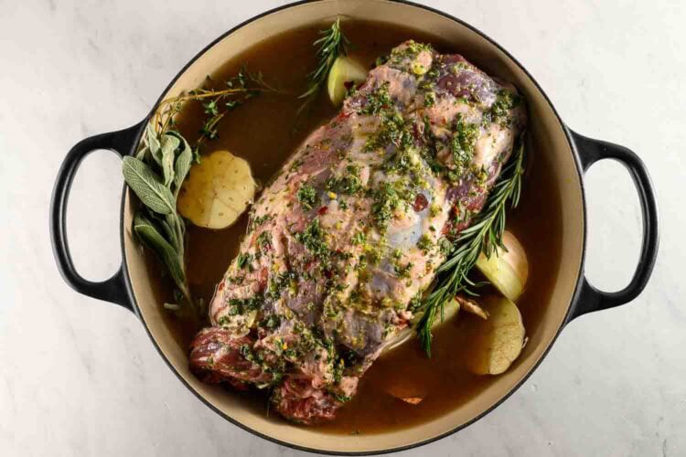 A seasoned Braised Leg of Lamb sits in a Dutch oven with fresh herbs, onions, and broth, ready to be cooked.