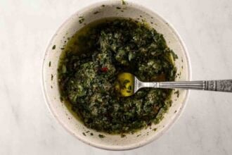 A bowl of chimichurri sauce with chopped herbs and olive oil, perfect for drizzling over Oven Braised Leg of Lamb, with a spoon resting inside the bowl on a light-colored surface.