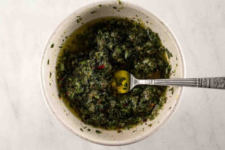 A bowl of chimichurri sauce with chopped herbs and olive oil, perfect for drizzling over Oven Braised Leg of Lamb, with a spoon resting inside the bowl on a light-colored surface.