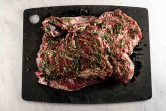 Raw marinated steak with herbs and oil on a black cutting board placed on a white surface, perfect for pairing with an oven braised leg of lamb for a hearty meal.