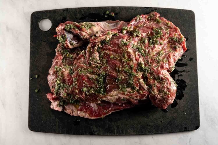 Raw marinated steak with herbs and oil on a black cutting board placed on a white surface, perfect for pairing with an oven braised leg of lamb for a hearty meal.
