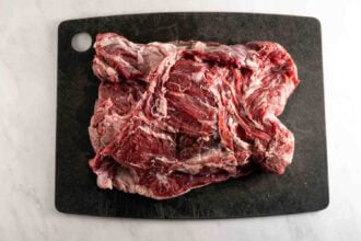 A slab of raw beef sits on a black cutting board atop a white surface, ready to be transformed into a succulent oven braised leg of lamb or an aromatic oven roasted lamb feast.