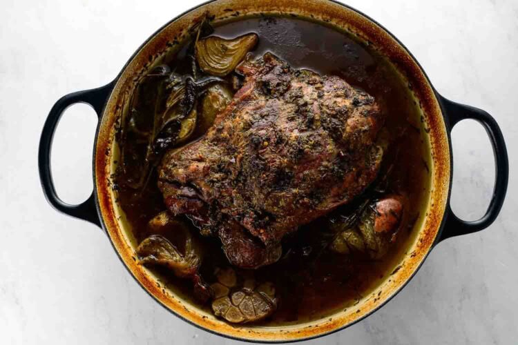 A braised leg of lamb with herbs, onions, and garlic sits in a black cast iron Dutch oven filled with savory broth.