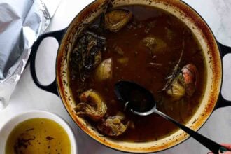 A large pot filled with cooked vegetables and broth for braised leg of lamb; a black spoon rests inside. A bowl of strained broth sits nearby on a white surface.