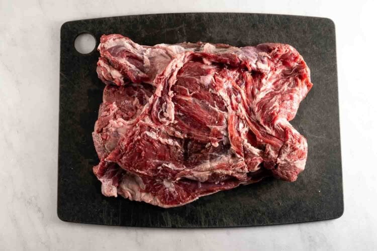 A slab of raw beef sits on a black cutting board atop a white surface, ready to be transformed into a succulent oven braised leg of lamb or an aromatic oven roasted lamb feast.