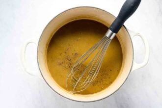 A metal whisk rests in a white pot filled with brown gravy, sprinkled with black pepper, beside an oven braised leg of lamb on a white surface.