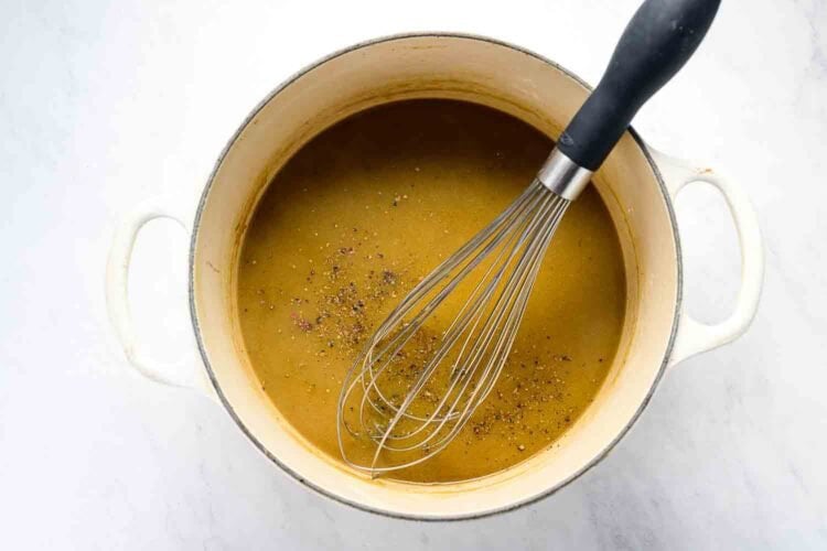 A metal whisk rests in a white pot filled with brown gravy, sprinkled with black pepper, beside an oven braised leg of lamb on a white surface.
