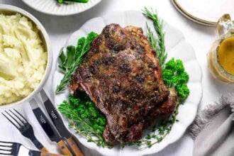 A braised leg of lamb garnished with fresh herbs on a white plate, served alongside mashed potatoes, savory gravy, and utensils atop a white table.
