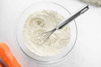 A glass bowl filled with flour mixture and a metal whisk, with two whole carrots placed nearby on a white surface—perfect for preparing delicious Carrot Fritters or other carrot recipes.