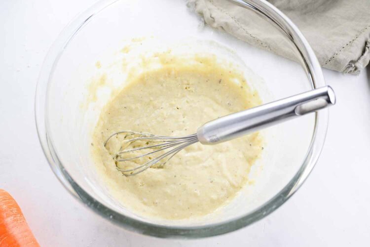 A metal whisk rests in a glass bowl filled with thick, creamy batter for carrot fritters on a white surface, with a beige cloth nearby.