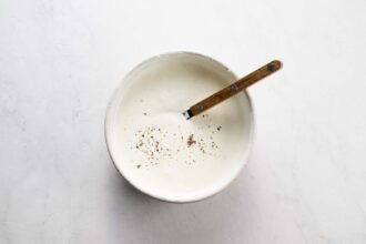 A bowl of creamy white sauce topped with cracked black pepper, with a wooden-handled spoon resting inside—perfect for dipping crispy carrot fritters or other tasty vegetable fritters.