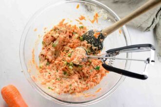 A glass bowl contains a mixture for Carrot Fritters, with grated carrots, chopped vegetables, and batter. A spoon and cookie scoop rest inside, while a carrot and beige napkin are placed nearby.