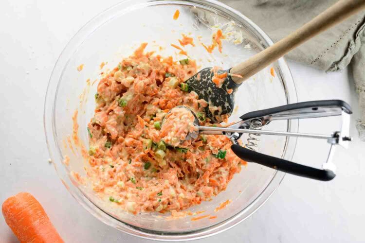 A glass bowl contains a mixture for Carrot Fritters, with grated carrots, chopped vegetables, and batter. A spoon and cookie scoop rest inside, while a carrot and beige napkin are placed nearby.