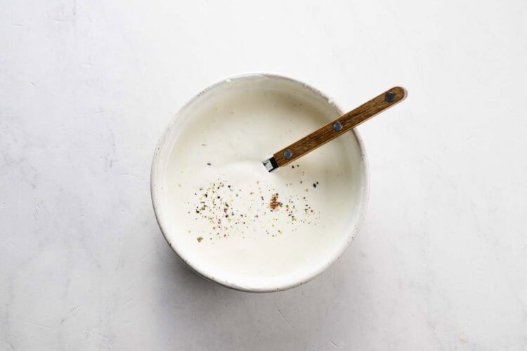 A bowl of creamy white sauce topped with cracked black pepper, with a wooden-handled spoon resting inside—perfect for dipping crispy carrot fritters or other tasty vegetable fritters.