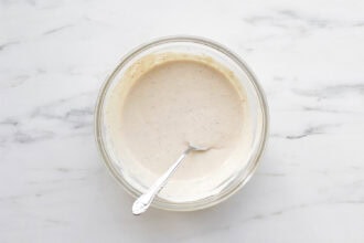 A glass bowl filled with creamy Caesar Salad Dressing made with Greek Yogurt, with a spoon resting inside, placed on a white marble surface.