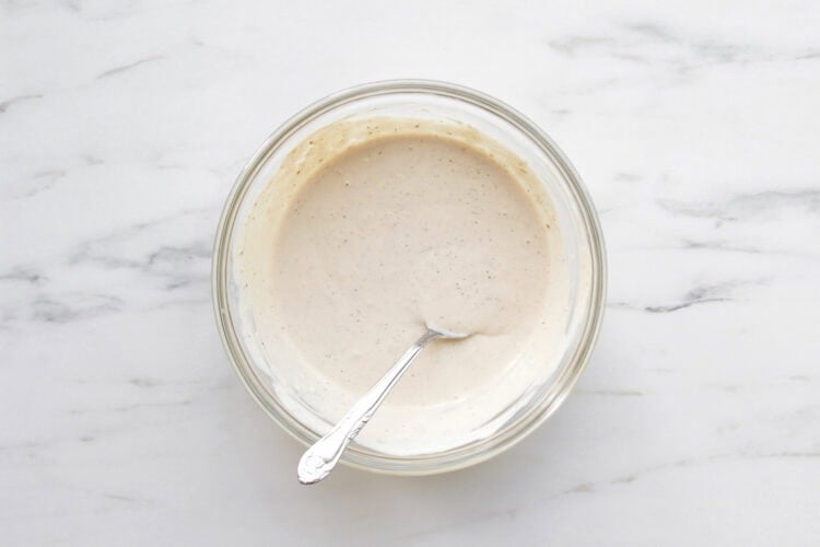 A glass bowl filled with creamy Caesar Salad Dressing made with Greek Yogurt, with a spoon resting inside, placed on a white marble surface.