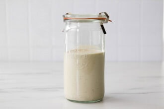 A glass jar filled with a beige, creamy Greek Yogurt Caesar Salad Dressing sits on a white surface against a tiled white background, promising rich flavor in every spoonful.