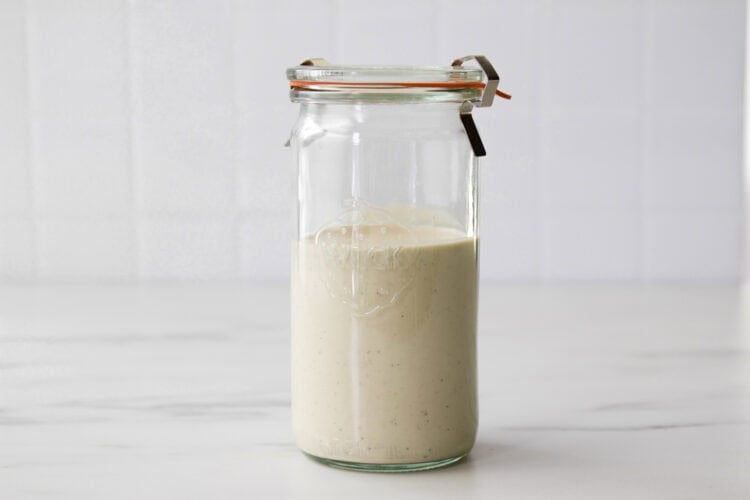 A glass jar filled with a beige, creamy Greek Yogurt Caesar Salad Dressing sits on a white surface against a tiled white background, promising rich flavor in every spoonful.