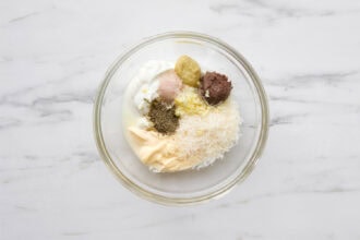 A glass bowl containing Caesar Salad Dressing made with mayonnaise, grated cheese, ground pepper, salt, mustard, minced garlic, anchovy paste, and Greek Yogurt on a white marble surface.