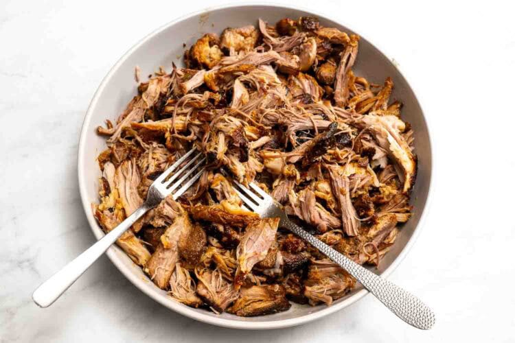 A bowl of Hot Honey Pulled Pork with two forks resting inside, placed on a white surface.
