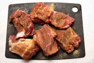 Six pieces of raw pork coated with dry seasoning, perfect for making hot honey pulled pork, are arranged on a black cutting board.