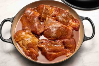Raw, seasoned pieces of meat are arranged in a round, enameled cast iron pot with a light layer of liquid at the bottom, ready to transform into flavorful Hot Honey Pulled Pork.