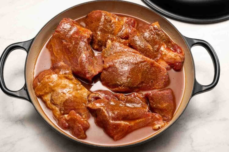 Raw, seasoned pieces of meat are arranged in a round, enameled cast iron pot with a light layer of liquid at the bottom, ready to transform into flavorful Hot Honey Pulled Pork.
