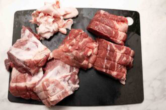 Raw pork cuts and chunks of fat arranged on a black cutting board, ready to be transformed into tender Hot Honey Pulled Pork on a white surface.