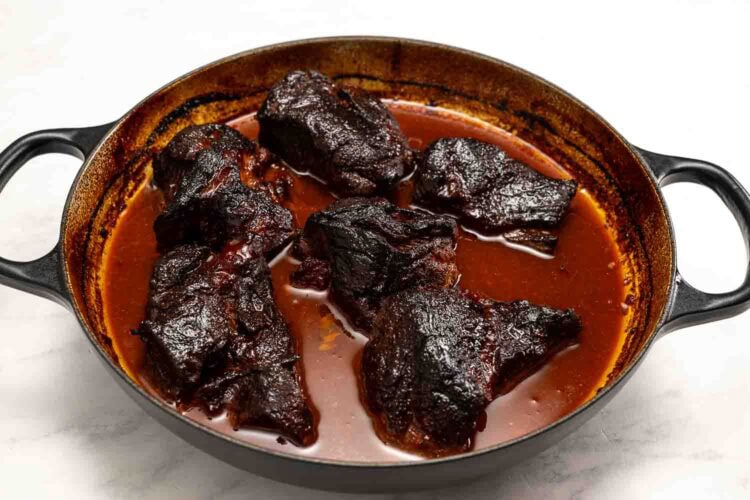 A black cast iron pot filled with Hot Honey Pulled Pork, featuring several large pieces of braised meat in a thick, dark reddish-brown sauce.