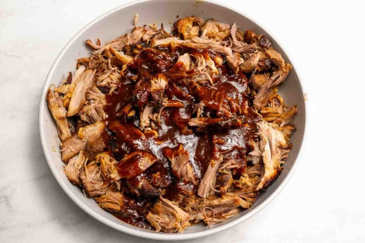 A bowl of hot honey pulled pork topped with barbecue sauce, placed on a white surface.