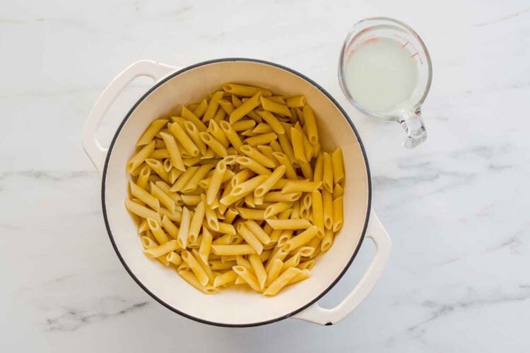 A white pot filled with cooked penne pasta sits on a marble surface next to a glass measuring cup containing pasta water—perfect for tossing with a creamy ricotta pasta sauce or topping with lemony shrimp.