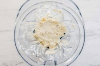 A blender bowl containing a thick, creamy white mixture—perfect for a luscious ricotta pasta sauce—sits on a white marble surface.