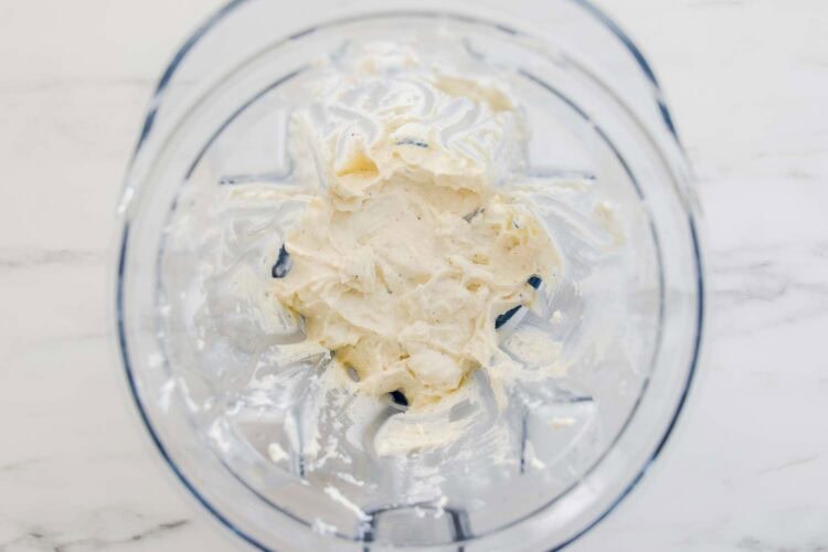 A blender bowl containing a thick, creamy white mixture—perfect for a luscious ricotta pasta sauce—sits on a white marble surface.