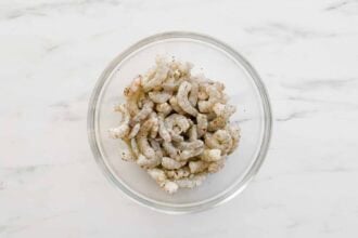 A glass bowl filled with raw shrimp pieces seasoned with spices, perfect for adding to a lemony pasta or tossing into a creamy ricotta pasta sauce, placed on a white marble surface.