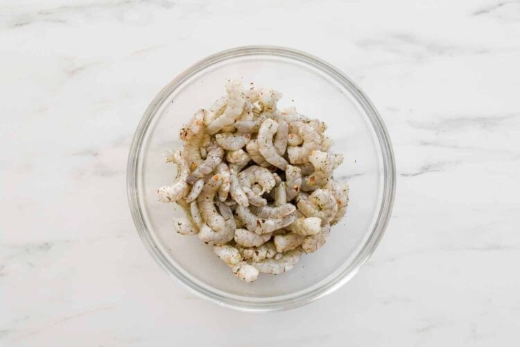 A glass bowl filled with raw shrimp pieces seasoned with spices, perfect for adding to a lemony pasta or tossing into a creamy ricotta pasta sauce, placed on a white marble surface.
