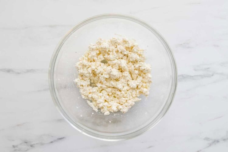 A clear glass bowl containing crumbled white cheese for a Lemony Ricotta Pasta Sauce sits on a white marble surface.