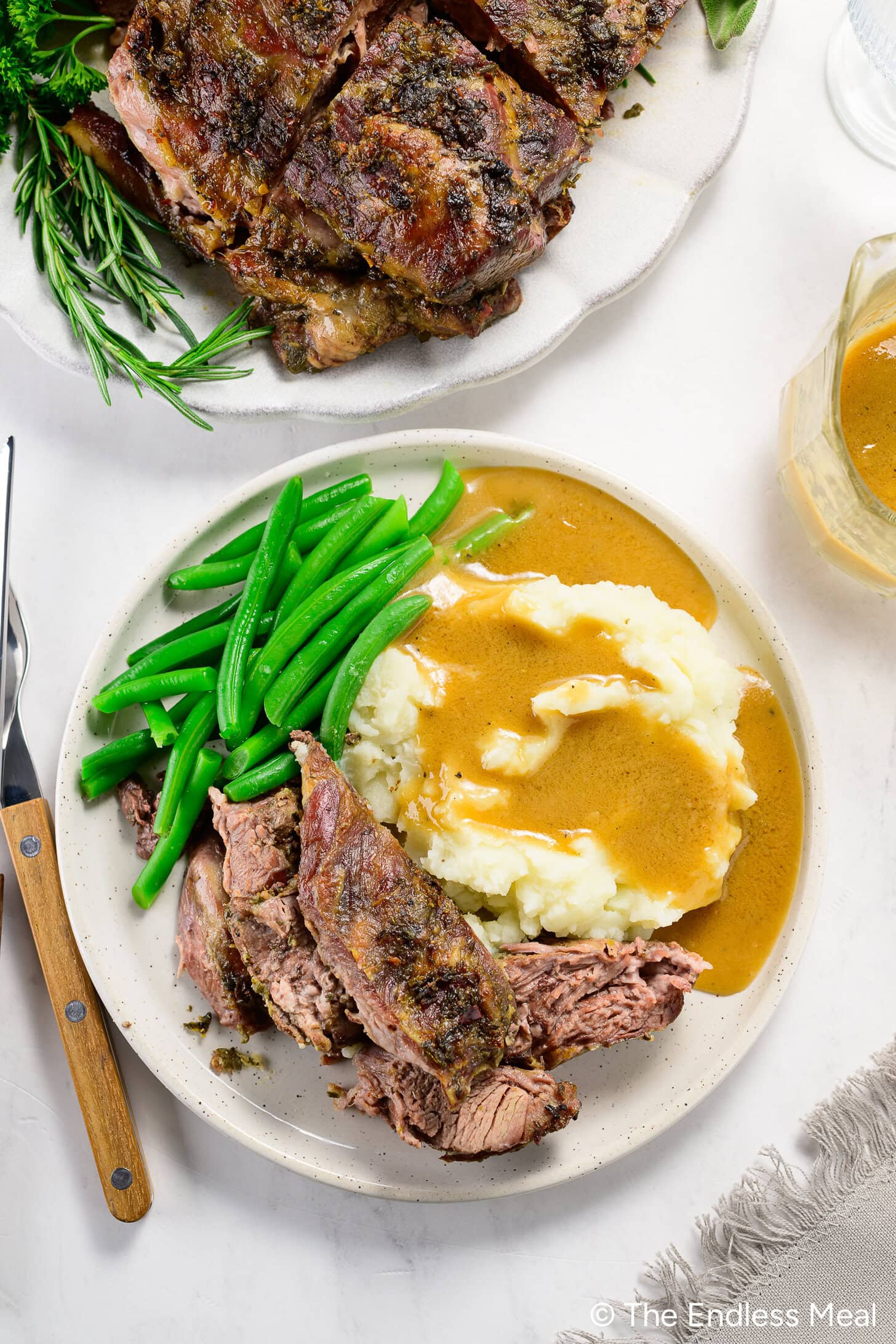 Plate with sliced boneless leg of lamb, mashed potatoes with gravy, and green beans. A fork is on the side. There is extra lamb, herbs, and gravy nearby.