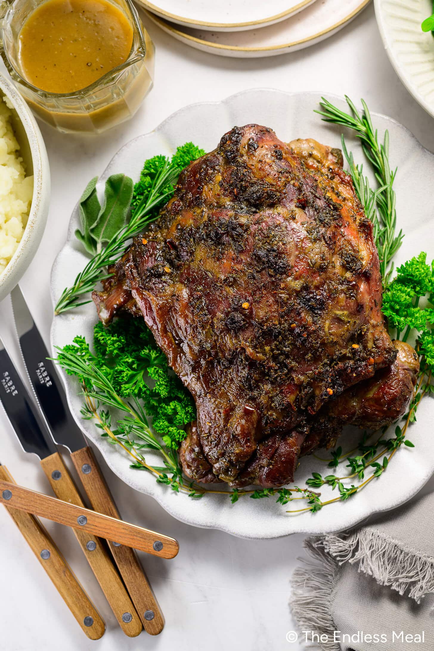 Roast lamb garnished with fresh herbs on a white plate, surrounded by side dishes, carving knives, and a bowl of gravy.