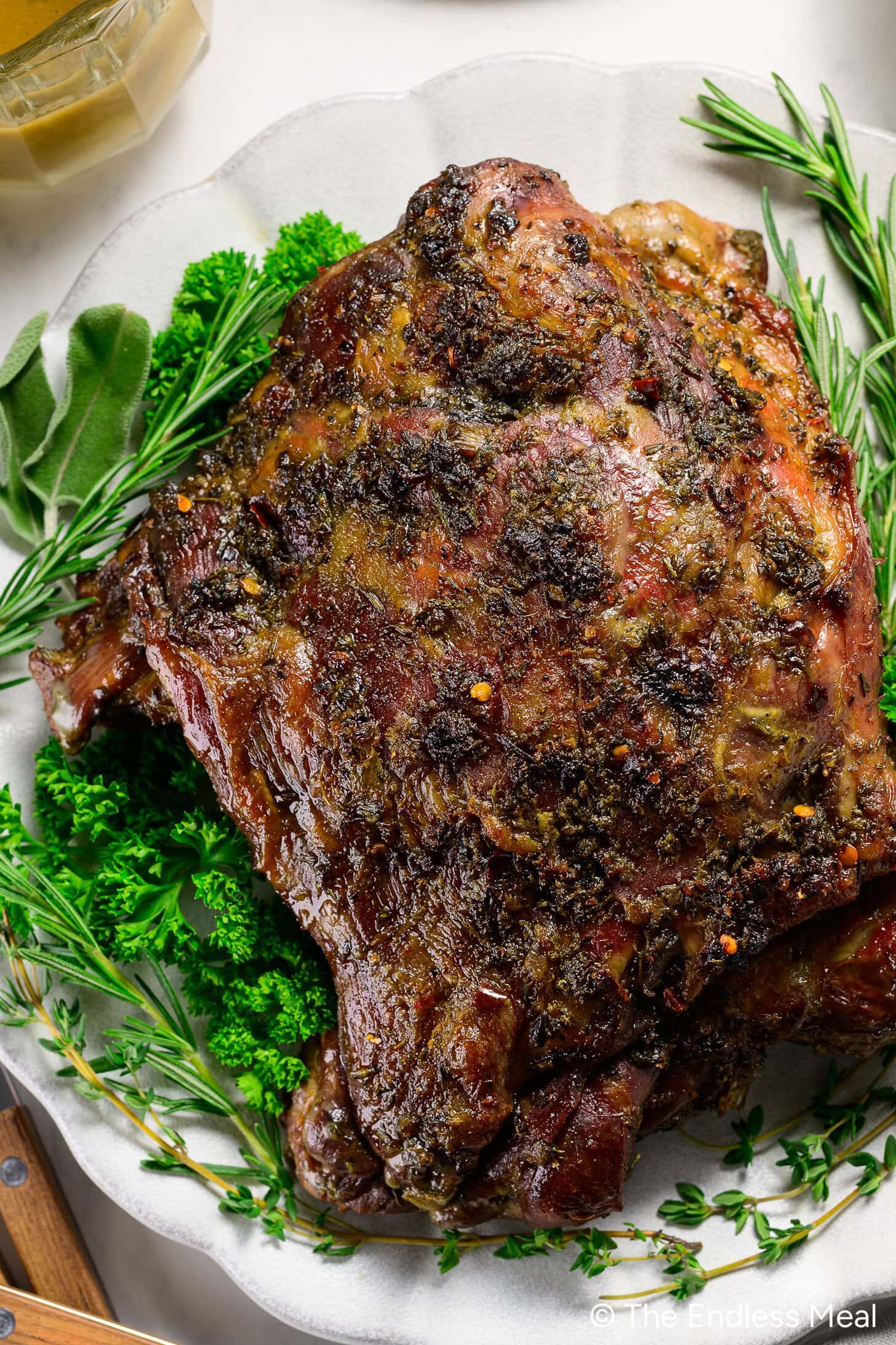 A cooked, herb-crusted boneless braised leg lamb roast sits on a white platter, garnished with fresh rosemary, parsley, and sage.