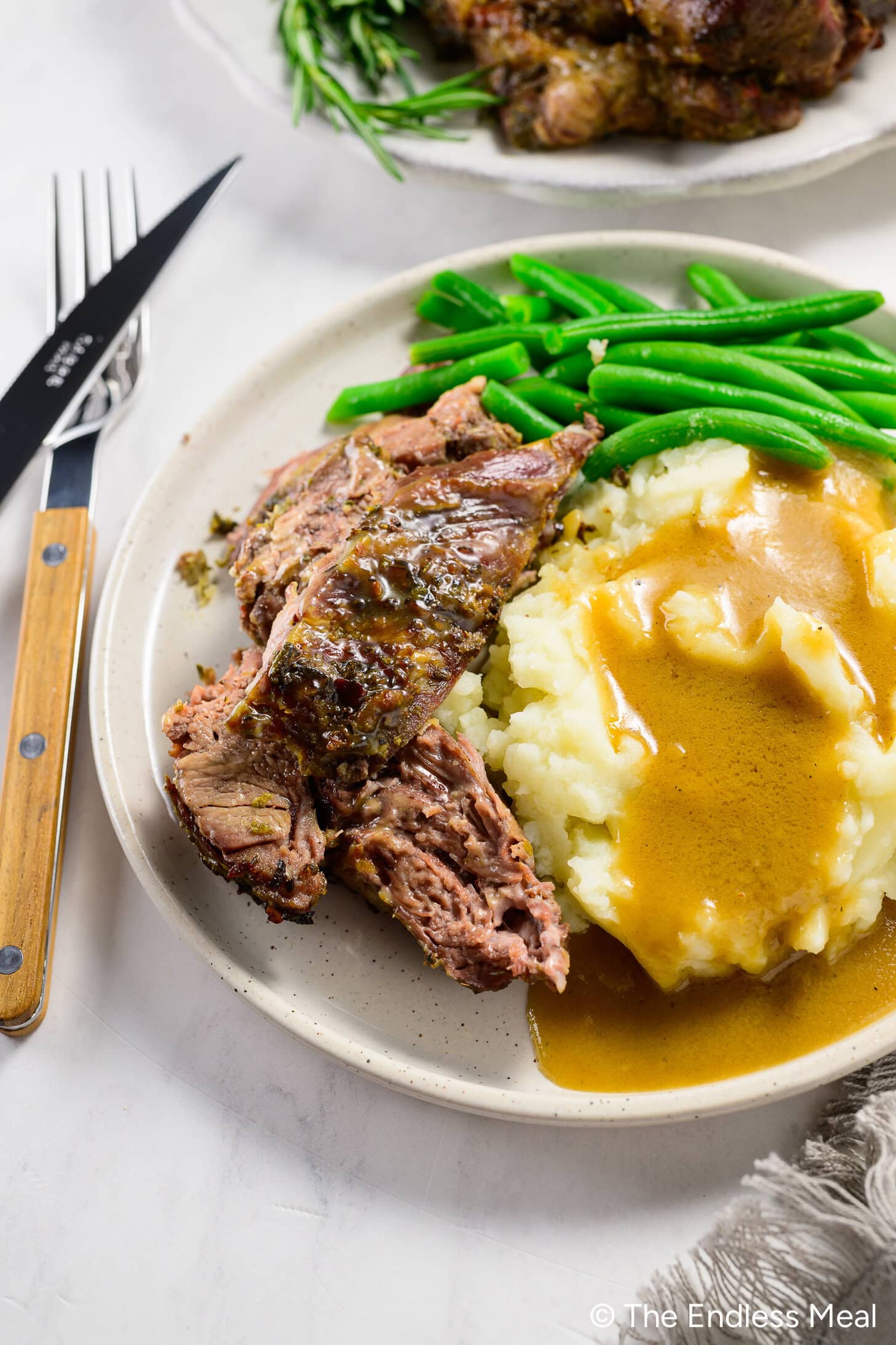 A plate with sliced roast beef, mashed potatoes topped with brown gravy, and green beans, next to a fork and knife on a white surface.