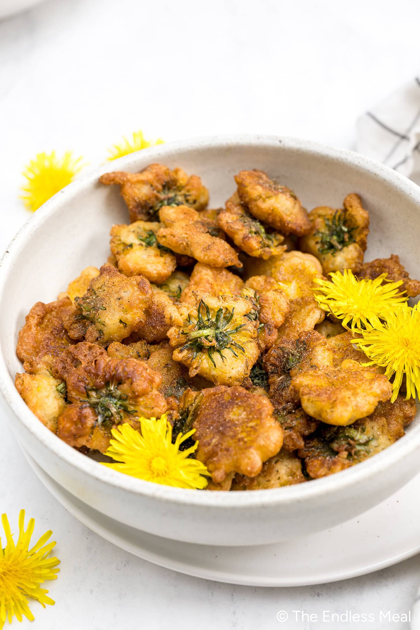 A white bowl filled with golden-brown Dandelion Donuts, garnished with fresh yellow dandelion flowers, on a white surface.