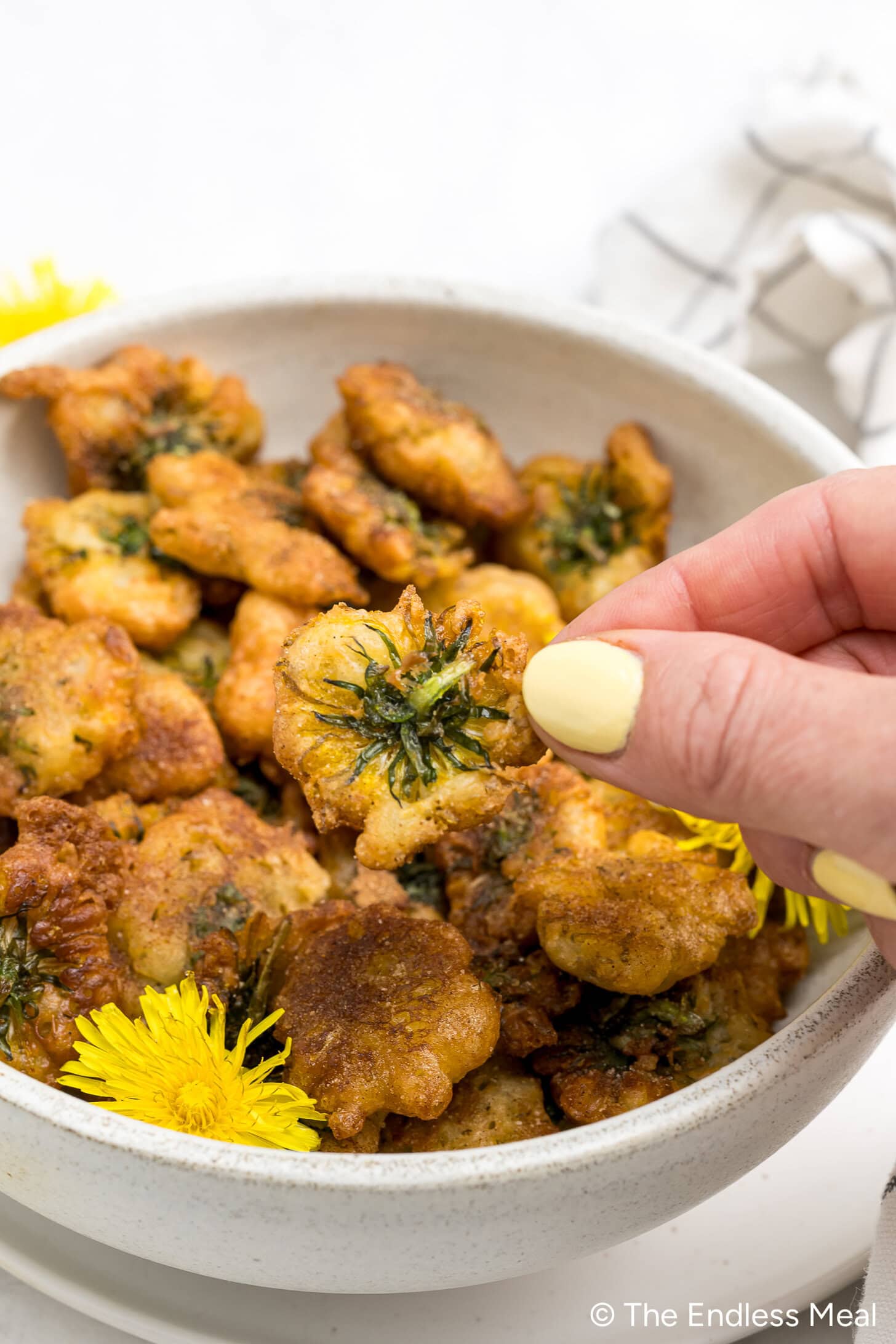 A hand holding a fried dandelion fritter—reminiscent of vegan donuts—above a bowl brimming with more fritters and fresh dandelion flowers.